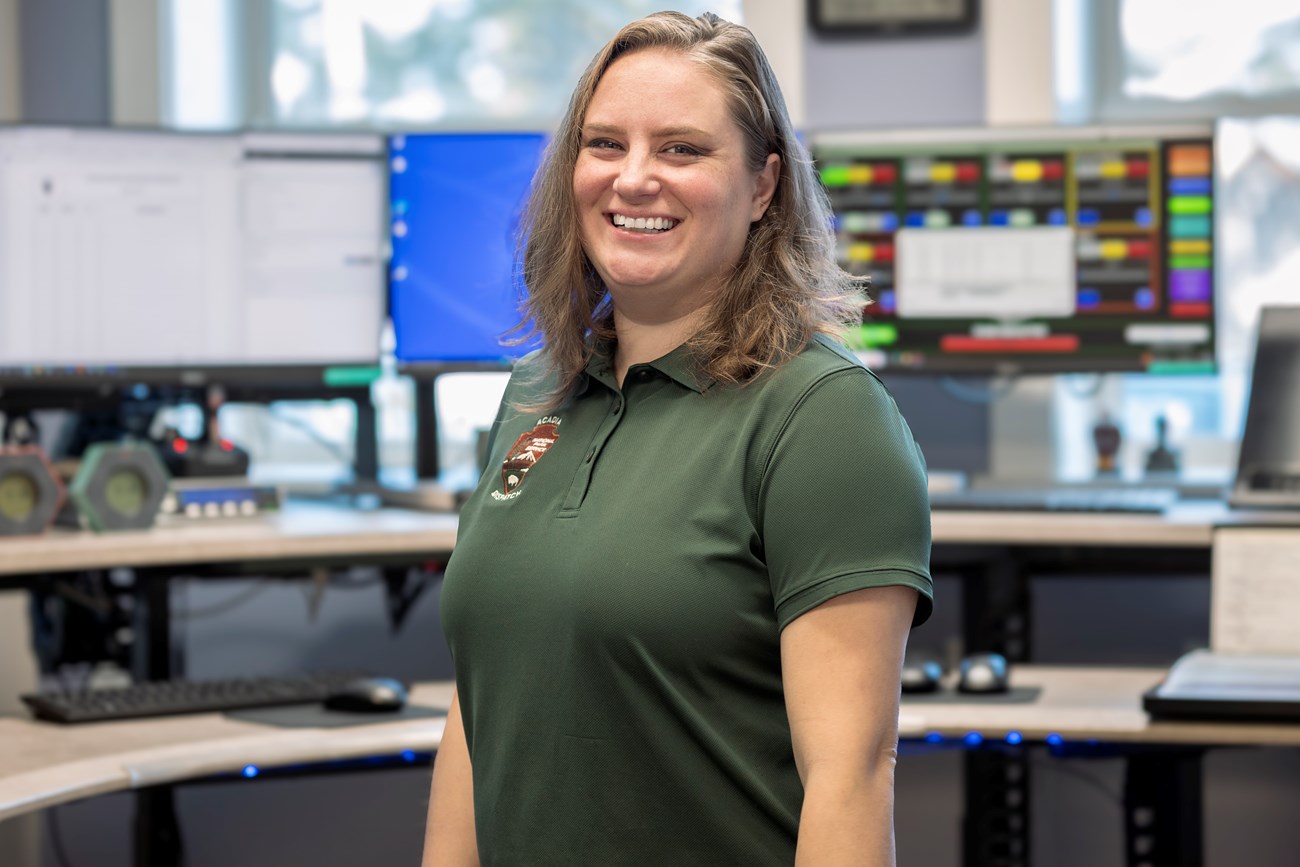 Brittany Ashcraft, Public Safety Officer, stands in front of the park’s Dispatch command center ready to receive any incoming calls and send any outgoing calls. Woman stands in front of five computer monitors at a dispatch center.