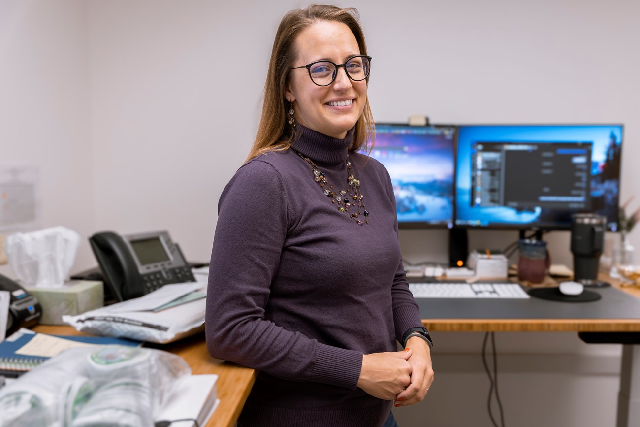 Stefanie Shattenberg, Recreation Fee Specialist, stands in front of her office desk. Woman stands in front of an office desk.