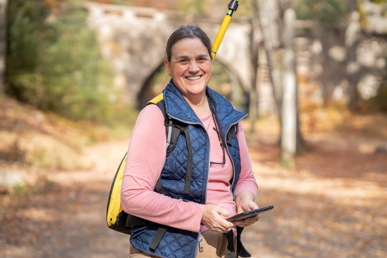 Karen Anderson, GIS Specialist, stands in front of a historic bridge on the carriage roads while using a mapping device. Woman with GNSS device stands in front of historic bridge on a bike path.