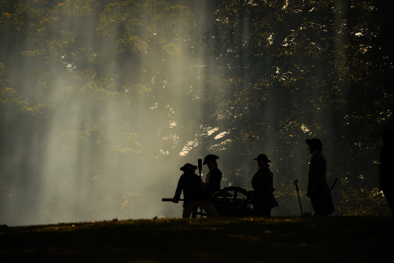 The silhouette of a cannon crew is illuminated by sunrays shining through smoke.
