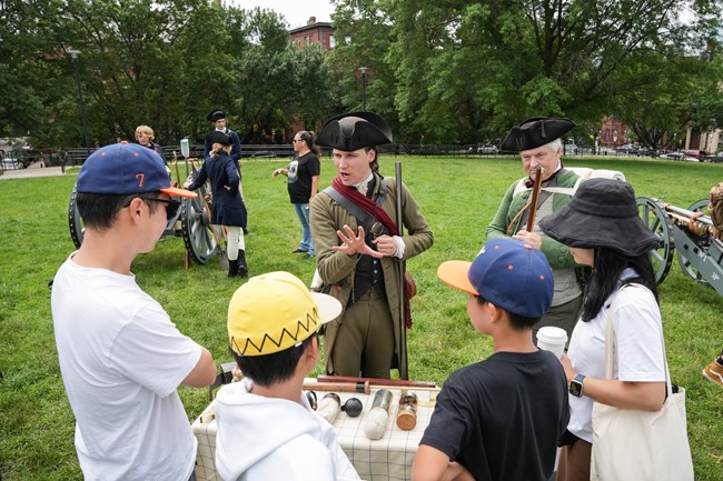 A ranger in 1770s militia kit speaks to a family who are looking at items on a table. Two cannon are in the background.