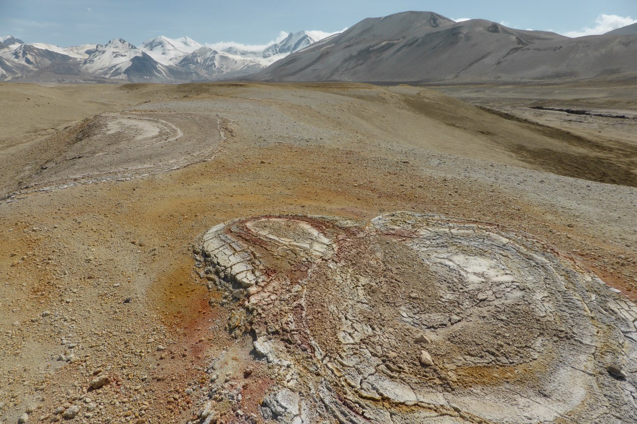 photo of a barren arctic landscape with oval features from extinct fumaroles on the surface. Mountains and snowy peaks are visible in the distance