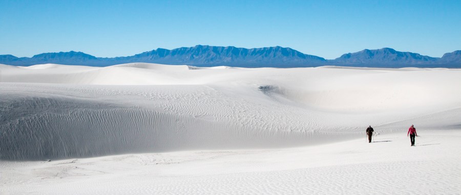 White Sands National Park landscape Two people in the distance hike on top of rolling pristine white sand dunes with blue mountains looming on the horizon.