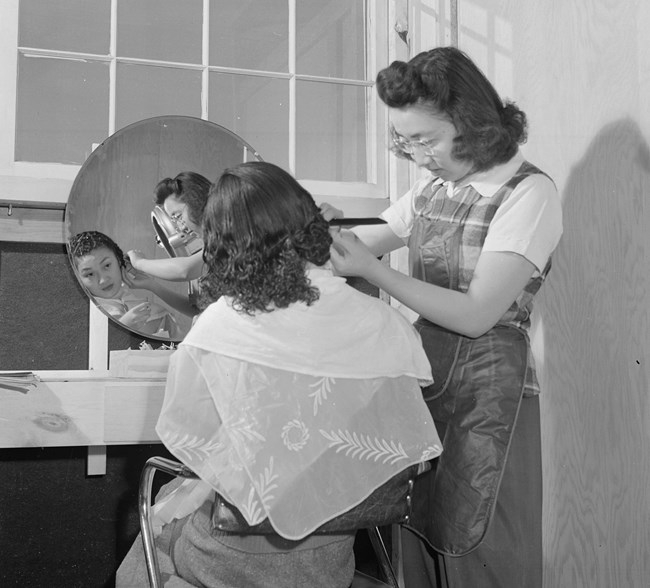Cooperative beauty salon A woman standing trims the hair of a seated Japanese American woman in a room with bare wooden walls
