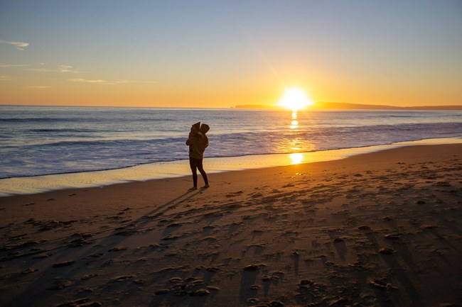 A mother and child watch the sunset at the water's edge on a sandy beach.