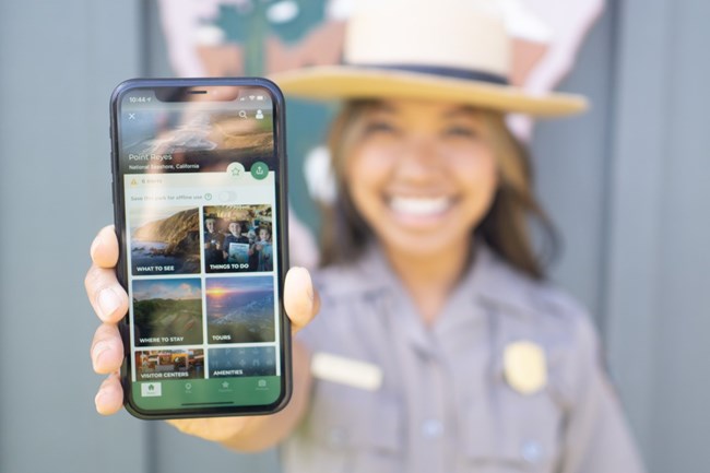 A female park ranger holds up a phone displaying the new NPS app!