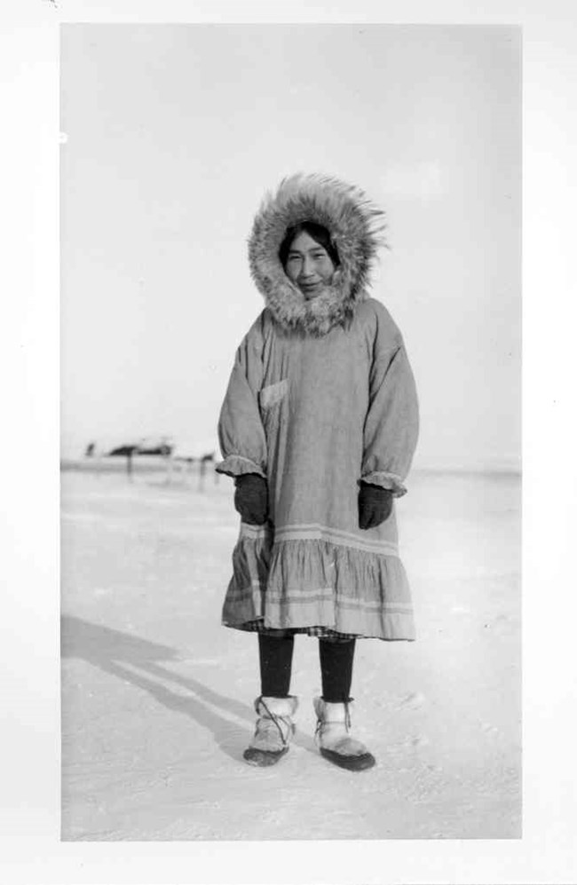Louise Tunmaqq, an Inupiat woman, is dressed in a parka with a large fur ruff, gloves, and traditional fur boots.