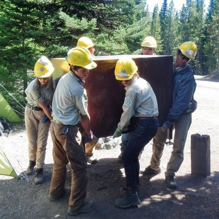 a group of young adults carrying a large metal food-storage box in a campground site