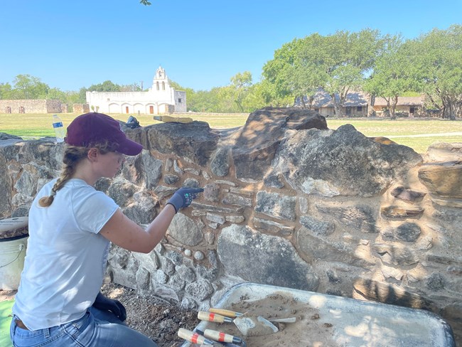 Woman repoints masonry wall. A white church is seen in the distance.