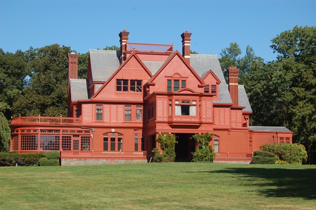 A large orange colored home with multiple chimneys sits on a grass lawn in front of trees.