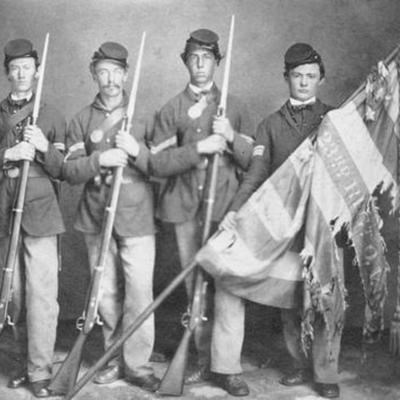 four young soldiers standing in a row wearing Civil War Union uniforms and one of them is holding a tattered American flag