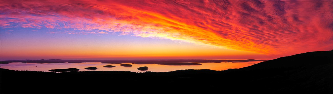 Panorama of sunrise with bright yellow, red, and orange clouds filling the sky at right, a water inlet with islands at center, and the silhouette of a mountain ridge framing the foreground Panorama of sunrise with bright yellow, red, and orange clouds filling the sky at right, a water inlet with islands at center, and the silhouette of a mountain ridge framing the foreground