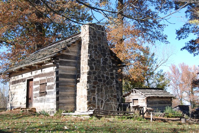 A log cabin with a stone chimney sits on a grassy field with trees in the background.  A second building is seen in the far right of the photo.