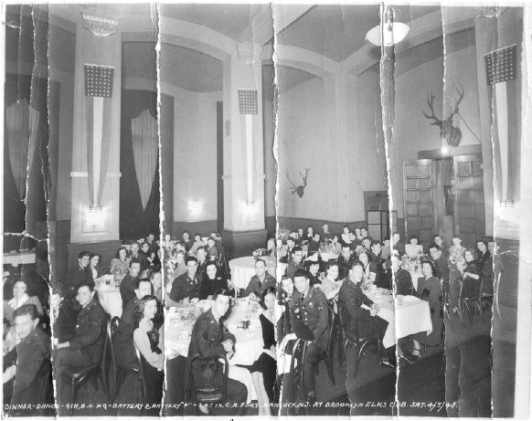 a large group of young soldiers and women sit around tabled in a social hall