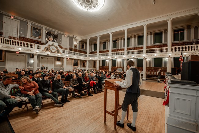Ranger in 1770s kit stands at a podium looking at a group of people sitting in the Great Hall of Faneuil Hall.