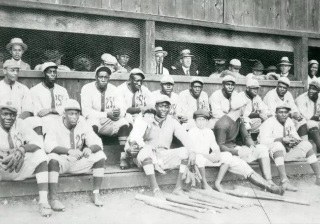 African American soldiers sitting in two rows in a dug out wearing baseball uniforms from the 1910s