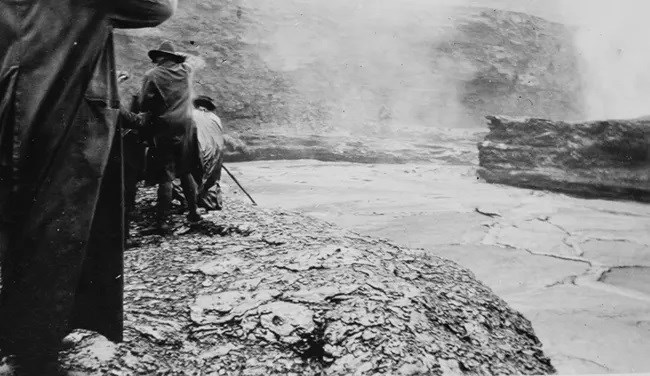 Soldiers take cover behind large rocks while an explosion goes off in the distance. The soldiers are making a hiking trail through heavy rock area.