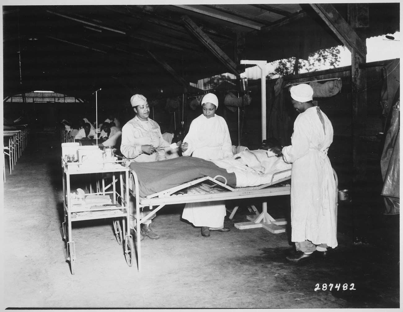 Black and white photo of three nurses in WWII uniform working around a Black man in a hospital bed.