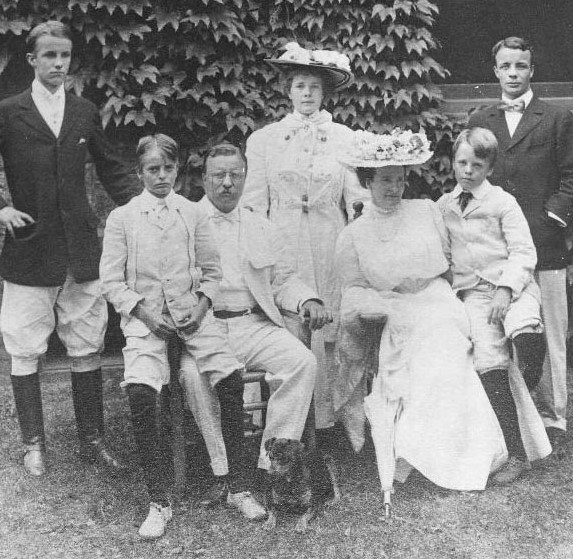 Roosevelt family seated and standing on a grassy lawn.  A tree and building is seen behind the family
