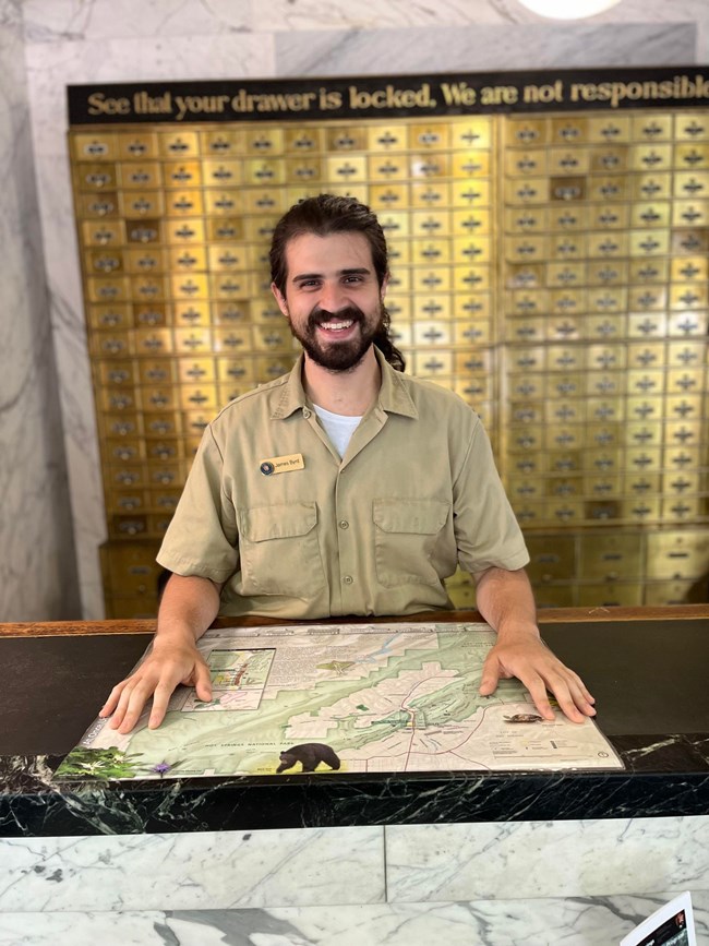 A young man with a dark beard and long dark hair wearing a khaki button-up shirt smiles from behind a desk.