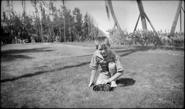 A black and white photo of Bobby Kennedy as a child petting a dark colored rabbit in a large lawn.