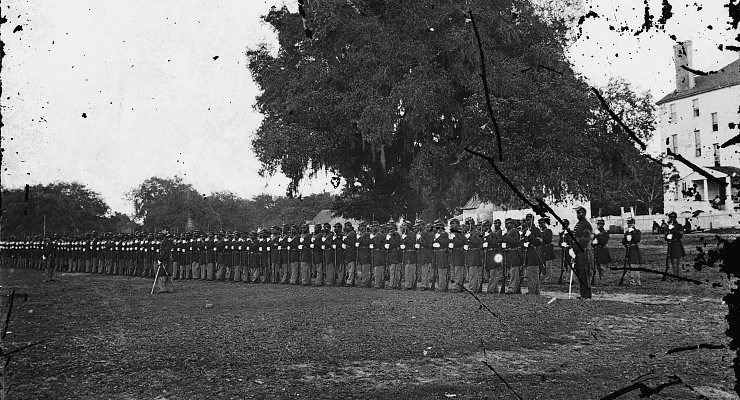 Long line of soldiers holding rifles standing in the lawn in front of a large white house.