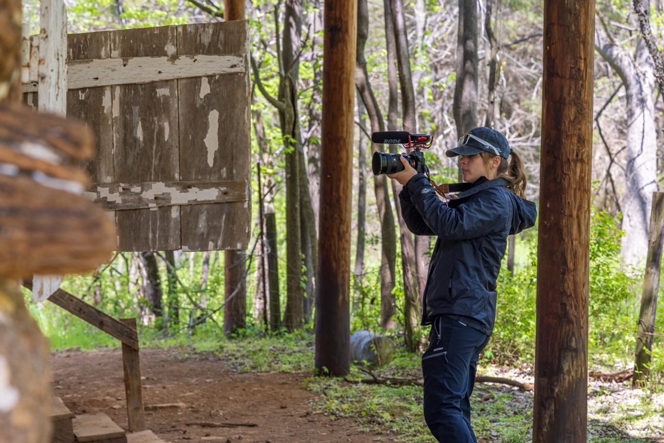 A person wearing a black jacket, black pants, and a baseball cap stands next to a log cabin holding a camera with a microphone attached to the top.
