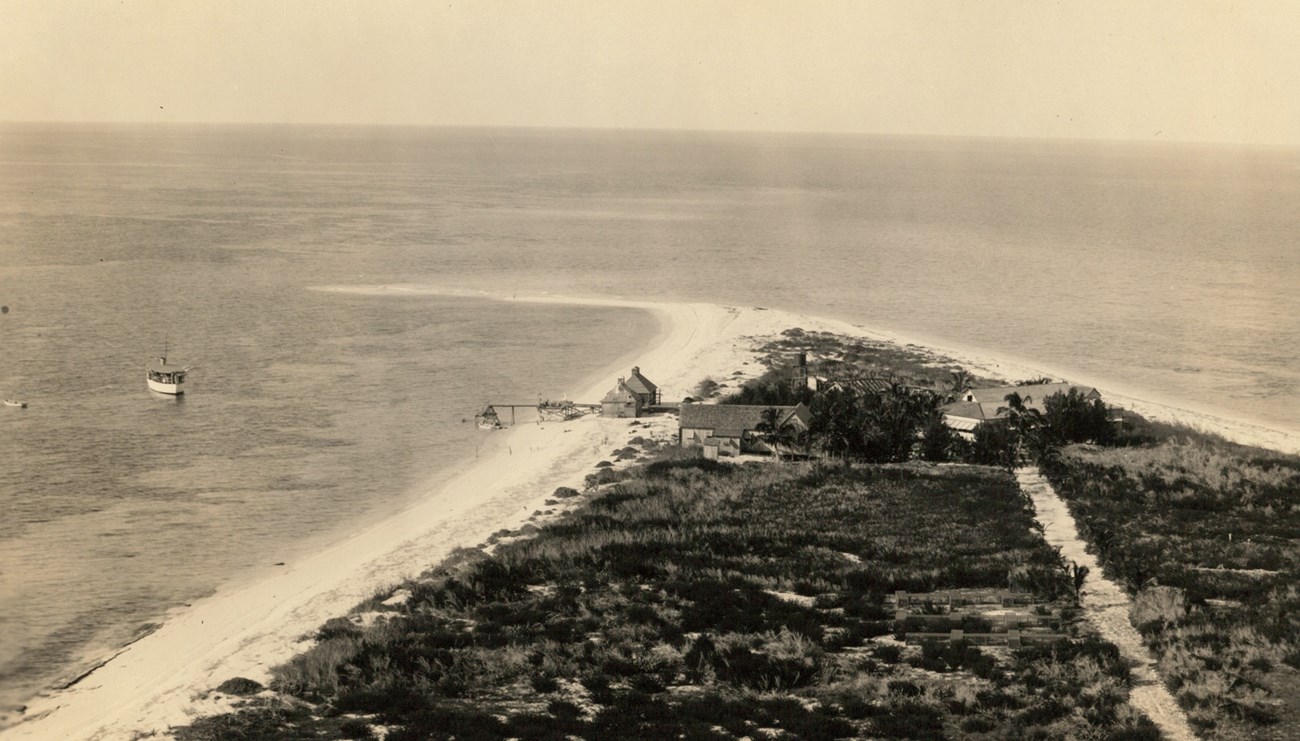 a hsitoric black and white photograph of a cluster of buildings on a sandy spit of land surrounded by ocean