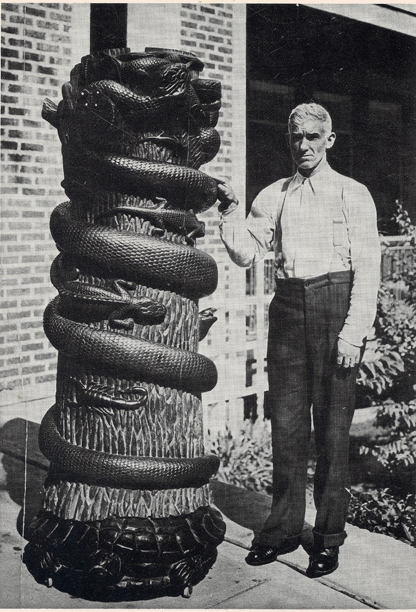 Man standing next to pole with carved snake encircling tree trunk. lizards and scorpions between snake coils.