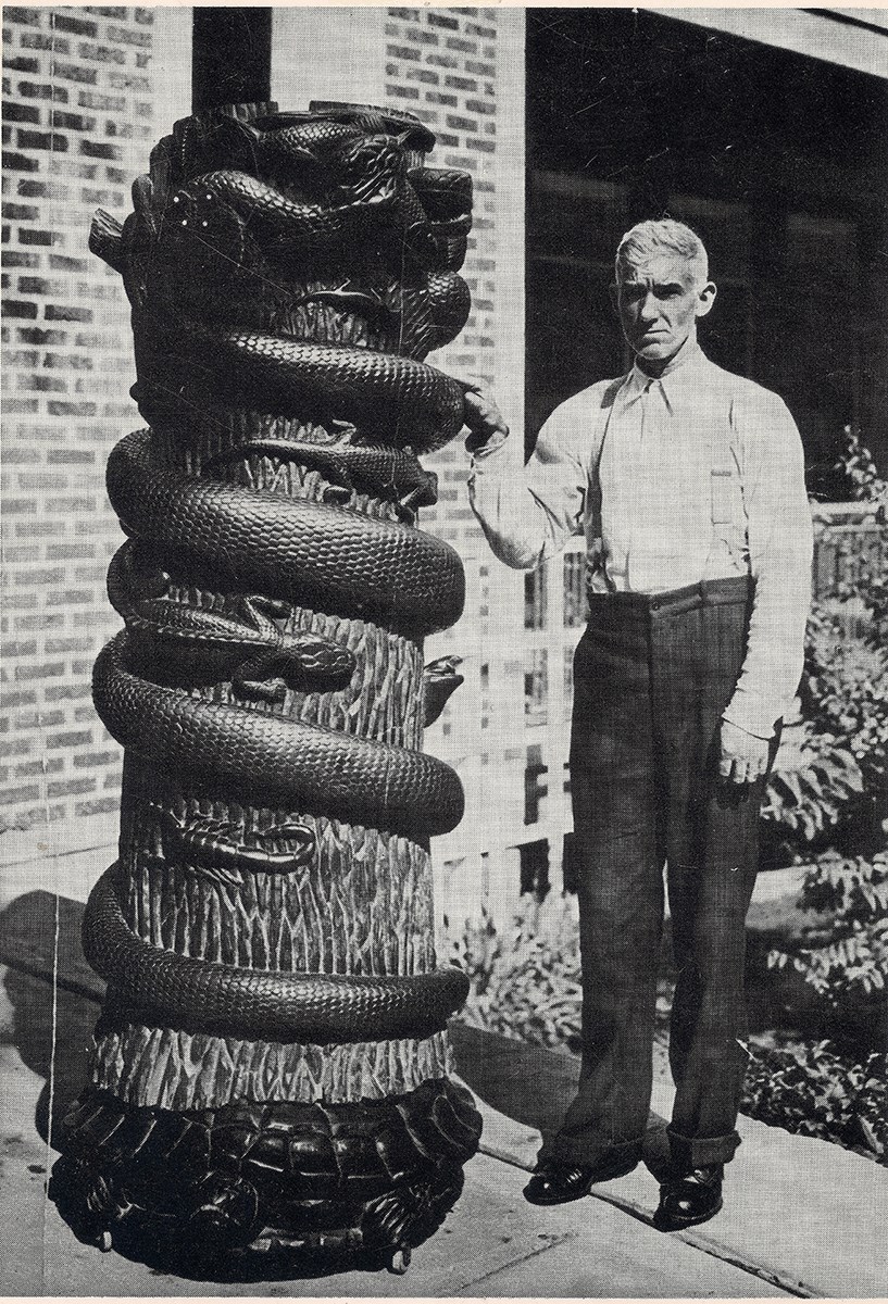 Tree Trunk Sculpture Man standing next to pole with carved snake encircling tree trunk. lizards and scorpions between snake coils.