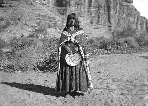 Havasupai woman Fannie Baanahmida In a black and white photo, a woman stands barefoot on the sand wearing historical Havasupai clothing.