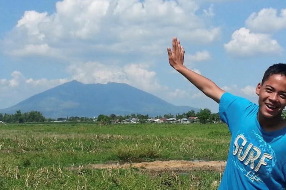 Rob and Mt. Arayat The author as a teenager wearing a blue "Surf Life" t-shirt, posing in an expansive grassy field with his arm extending up over a distant mountain with a distinct volcanic profile.