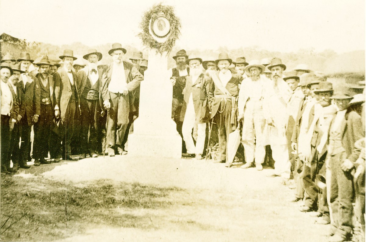 Sepia toned photograph of a group of men crowded around an obelisk shaped monument.