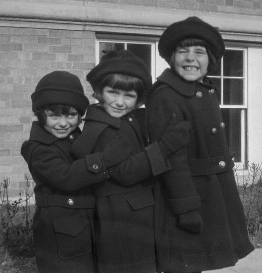 A black and white photo of three smiling girls in matching dark coats, hats, and bob haircuts standing in order of height, in front of a brick building. The two smaller girls each hold the arm of the girl in front. The tallest girl grins widely.