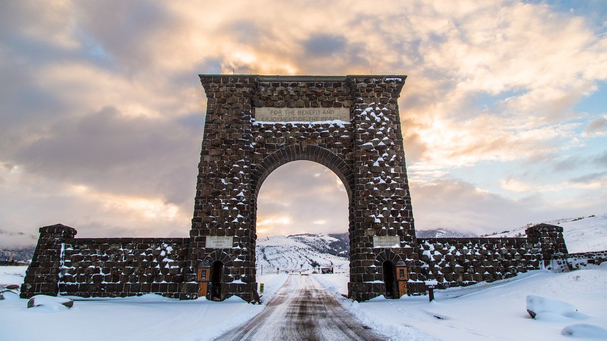 a snow-covered road that goes under a large stone archway at sunrise
