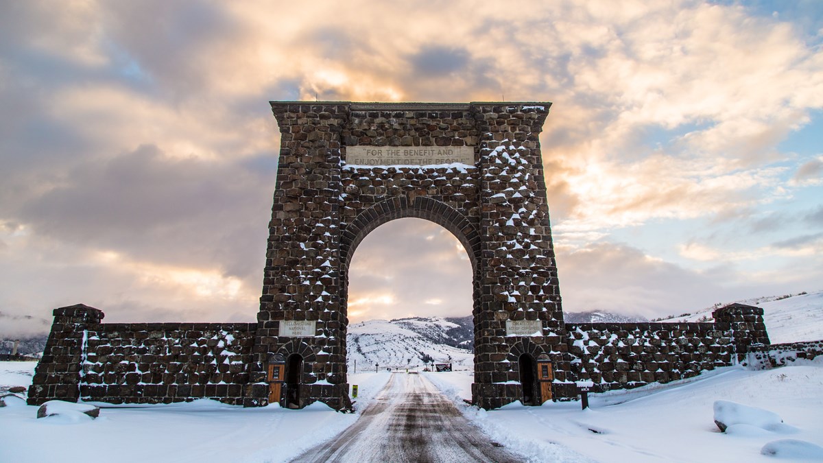 Winter Roosevelt Arch Sunrise a snow-covered road that goes under a large stone archway at sunrise