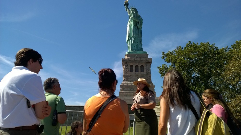 Several visitors listen as a park ranger talks about the history of the site, with the Statue of Liberty in the background.