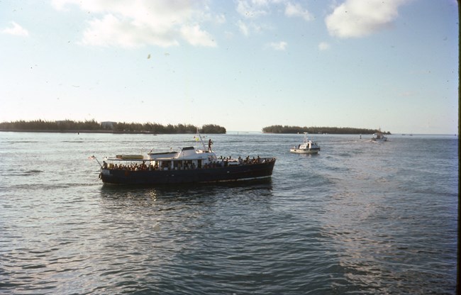 Boat arriving in Key West Harbor with Cuban refugees during the Mariel Boatlift. Two smaller boats are visible in the background. Two small islands covered with trees near the horizon line.