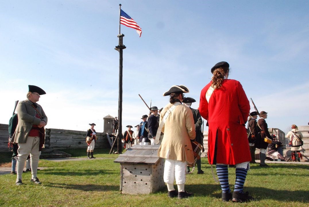 The American flag flies over the wall of the fort. Saluting it from underneath, a group of Continental Soldiers in stands formation.