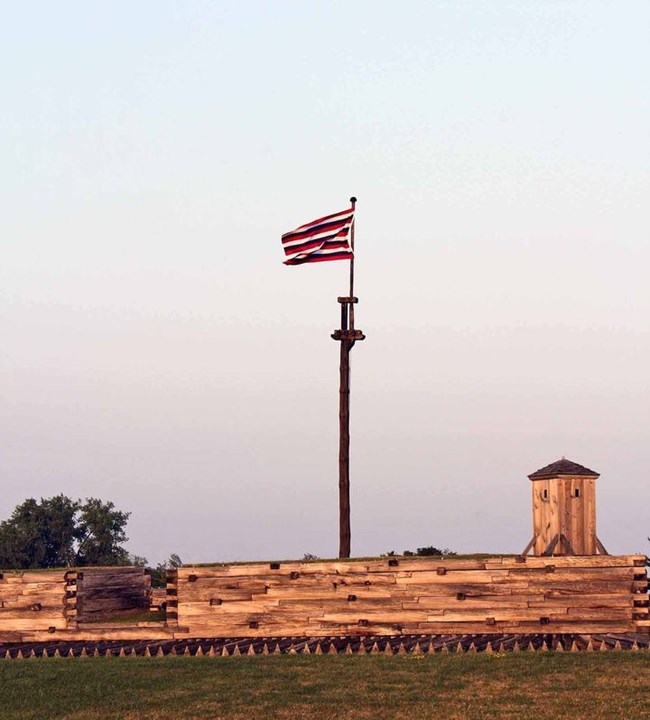 A flag on a wooden flag pole flies over the wall of the fort. It has alternating red, white, and blue horizontal stripes.