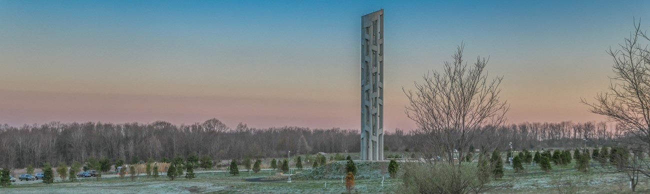 A tall narrow lattice-like tower memorial with a line of trees in the background.
