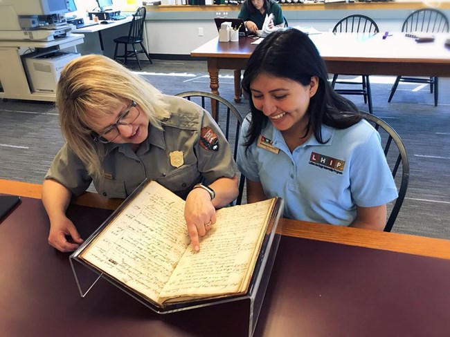 Maryann Zujewski looks over FRIENDSHIP OF SALEM documents with LHIP intern at the Philips Library park ranger looks over a book with another woman