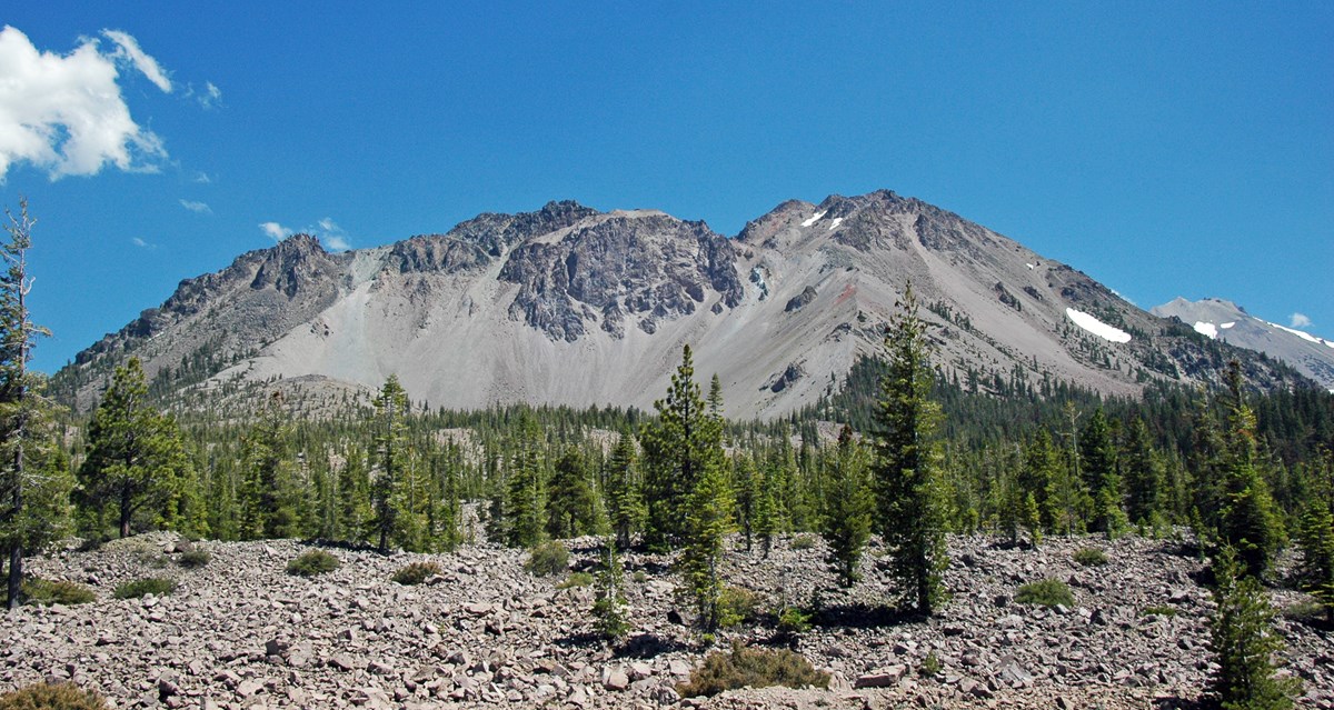 photo of a volcanic peak with trees at the base and a rocky debris field in the foreground
