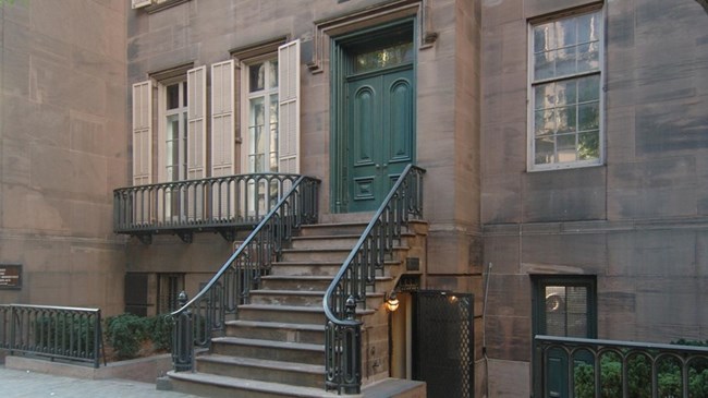 A four story stone building sits behind a sidewalk and a few trees.