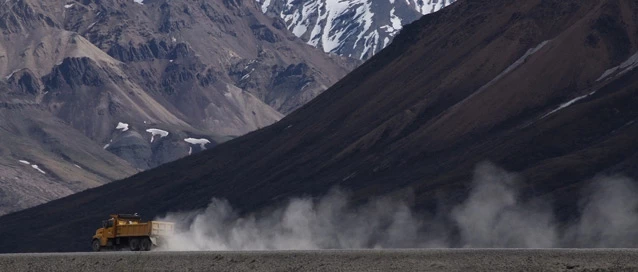A large orange maintenance truck drives down a gravel road past large rocky mountains, leaving a trail of dust behind it. A large orange maintenance truck drives down a gravel road past large rocky mountains, leaving a trail of dust behind it.