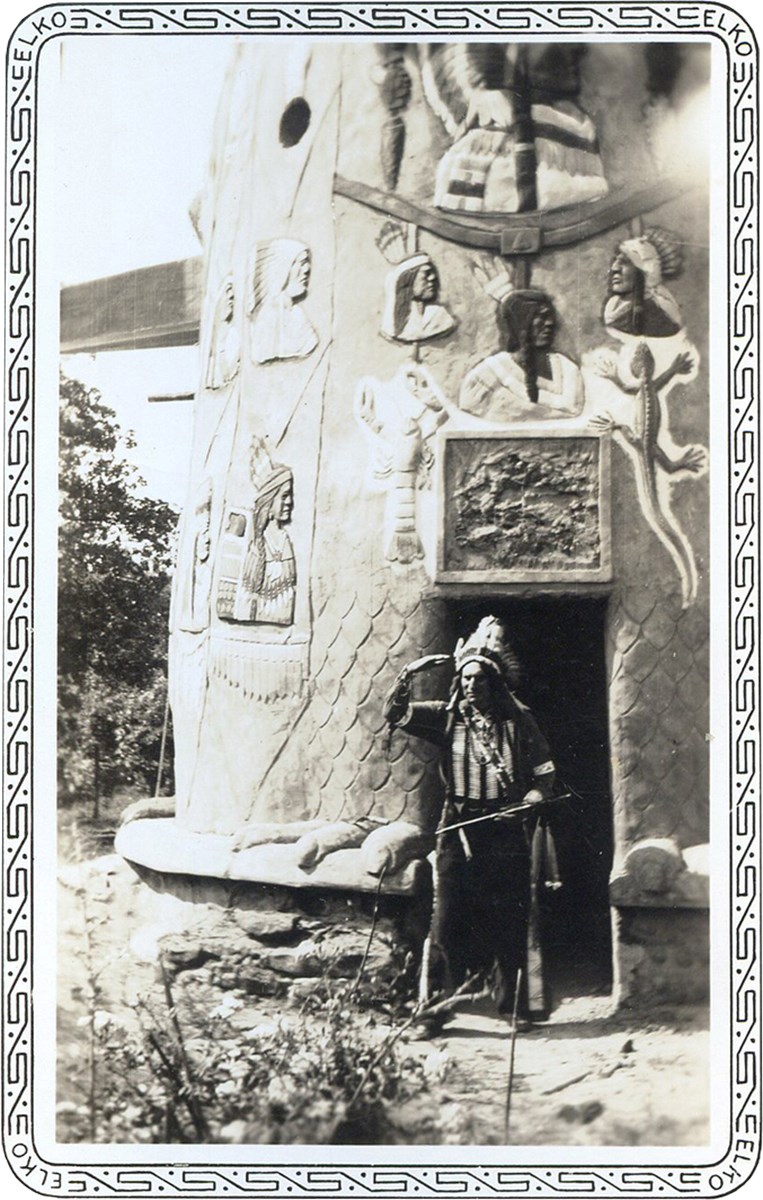 Man in Indian feather headdress and costume standing in doorway to totems with images of native Americans.