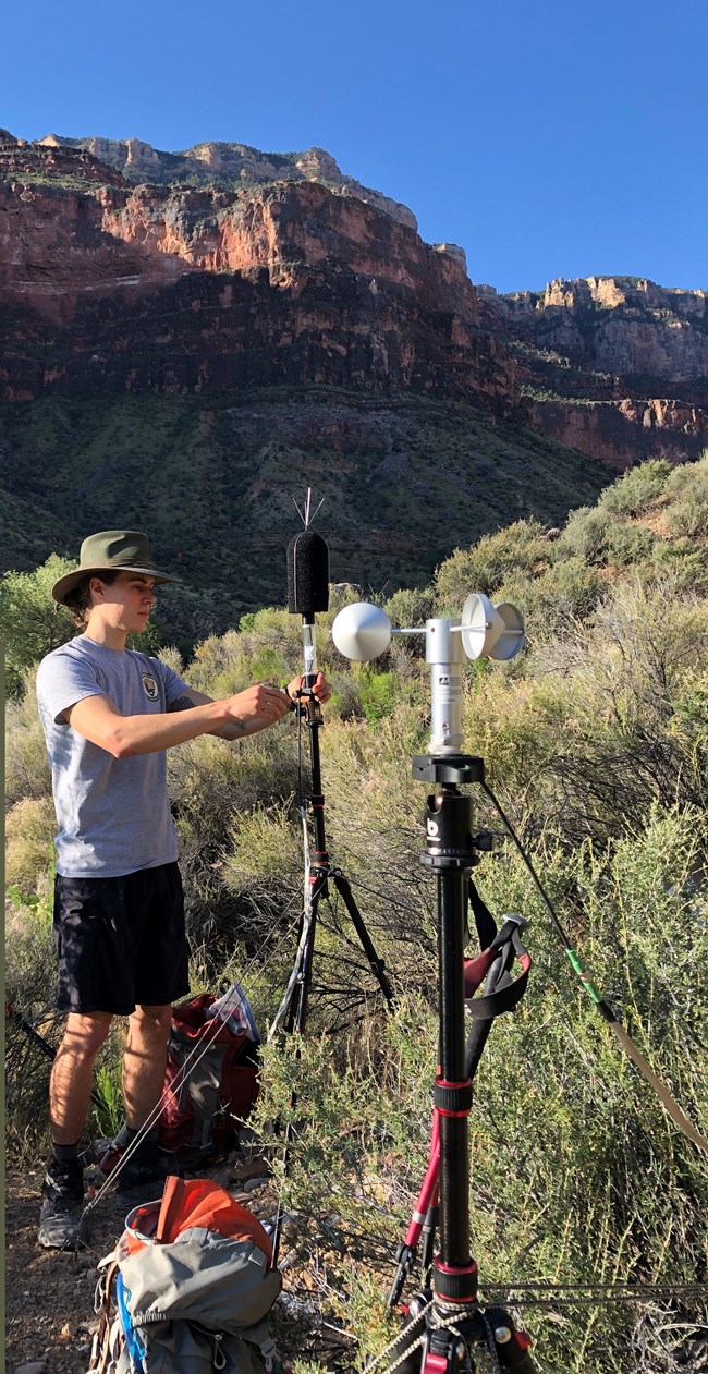 A man stands working with audio recording equipment on a tripod.