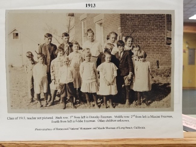 A black and white photo of the school children outside the brick school house.
