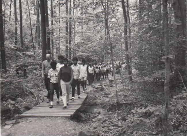 Summer in the Parks participants on a nature hike through the park. Most of the participants in the photograph are African American kids and teenagers, and they are walking across a footbridge in a heavily forested area.