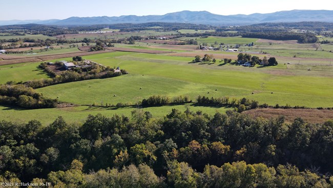 Piedmont Battlefield A bird’s eye view of green rolling hills, farms and farmland of the Shenandoah Valley. Rolls of hay dot the center field, and long shadows of trees, houses and barns suggest an early morning.